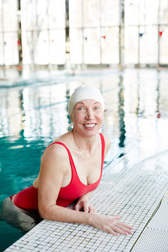 Cheerful mature female in red swimsuit and white rubber cap spending leisure in swimming pool