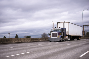 Big rig dark blue classic semi truck with dry van semi trailer running on wide highway