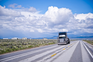 Black classic big rig semi truck transporting commercial cargo on flat bed semi trailer in the flat straight road in Nevada