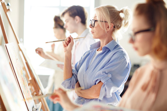 Pensive mature blond woman with paintbrush looking at her painting on easel