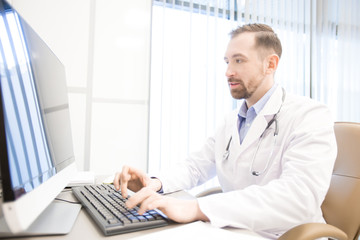 Young man in whitecoat sitting by computer monitor by his desk in clinics and consulting patients online
