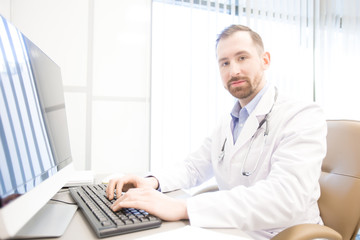 Young doctor looking at camera while sitting by computer monitor during online consultation