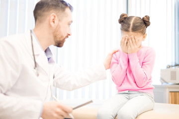 Fototapeta premium Male clinician comforting crying little girl during medical appointment in hospital