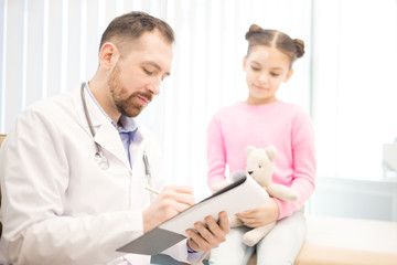 Young pediatrician writing down prescriptions for his little patient in modern clinics