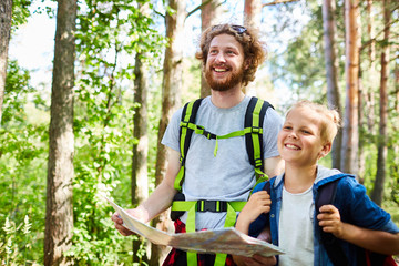 Cheerful backpackers with map looking for place to settle down while walking through forest on...