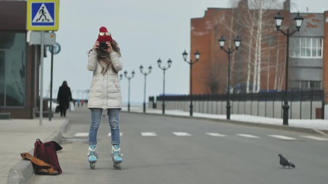 Young beautiful woman in red hat wearing sporty warm clothes and rollers, sitting on the asphalt road and taking pictures on a vintage camera