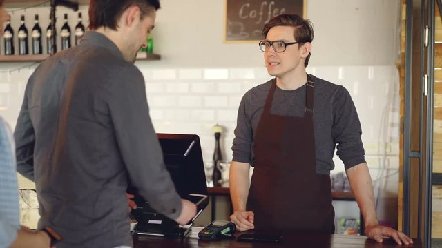 Friendly barista handsome man is selling takeout coffee and accepting contactless mobile payment with smart phone. Modern technolofy, coffee shop and banking concept.