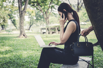 hand of thief picking up bag behind the woman, who sit and talk with friend on smartphone at the garden. concept of criminal, villain and bandit