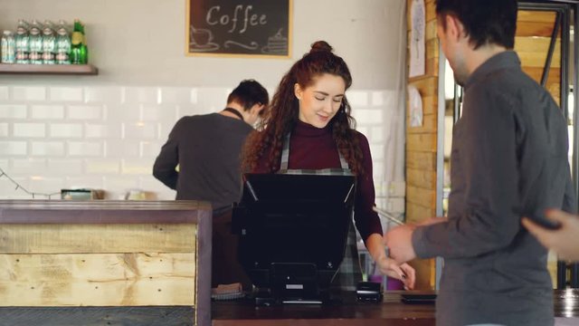 Attractive woman talkative cashier is accepting contactless payments with mobile phone and talking to customers and friendly waiter in apron is bringing takeaway coffee.