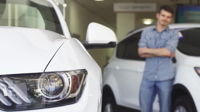 Selective focus on a car on the foreground copy space. Young cheerful man examining a car smiling happily. Male buyer choosing a new vehicle at the showroom. Retail, rental service concept.