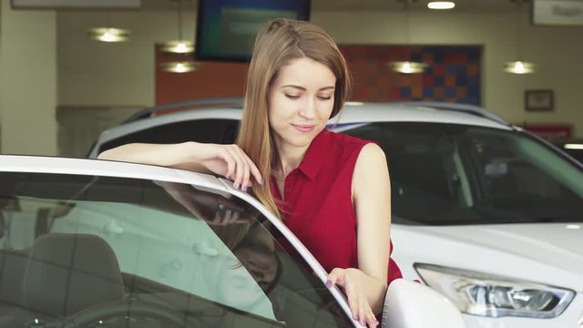 Attractive young female customer smiling joyfully touching gently a new convertible at the dealership salon. Beautiful woman buying a new automobile at the showroom. Consumerism, sales.