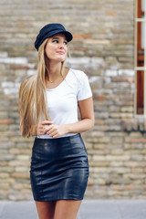 Young blonde woman wearing cap smiling near a brick wall.