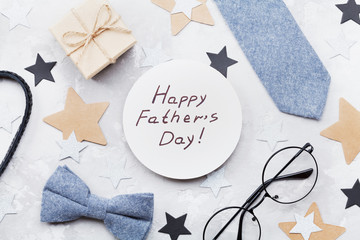 Happy Fathers Day card decorated bowtie, necktie, eyeglasses, gift box and stars on stone table top view in flat lay style.