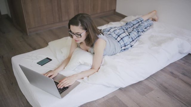 Pretty young woman in pajamas and glasses lying on soft mattress in stylish bedroom and browsing modern laptop in morning.