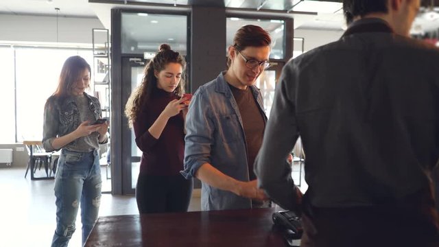 Cheerful customer in cafe is making payment with smartphone while queue of clients are waiting and using mobile phones. Takeaway drinks and modern banking concept.