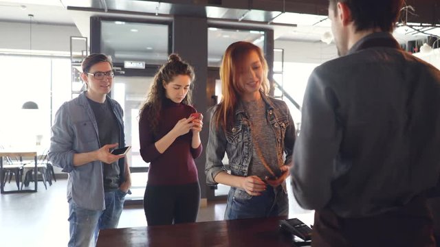 Queue of young people standing in coffee-house in the morning and buying takeaway drinks paying with cellphone. Modern lifestyle and contactless payment concept.