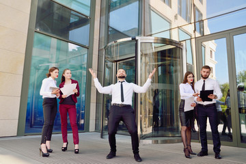 Fototapeta premium A young male businessman is enjoying a successful deal on the background of office workers. Demonstration of positive emotions and gestures. Career. Success. Shows a thumbs-up.