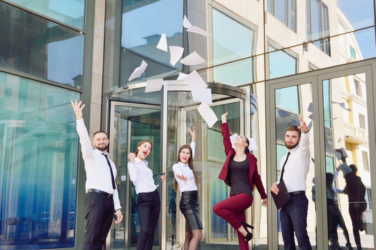 Office Workers Throw Paper Up Against The Background Of A Multi-storey Glass Office Building. Group Of Young People. Five Are Happy About The Successful Completion Of A Profitable Deal.