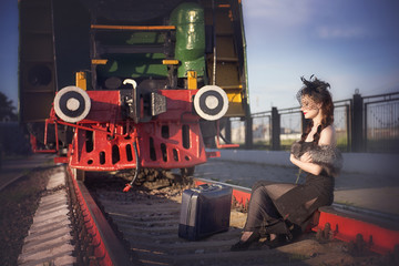 A woman in a vintage black dress sits on the rails near to a large suitcase against the backdrop of an old locomotive. Portrait of a fashionable woman in retro style.