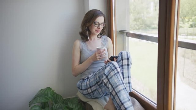 Attractive female in eyeglasses and pajamas sitting on window sill holding mug and using smartphone with earphones.