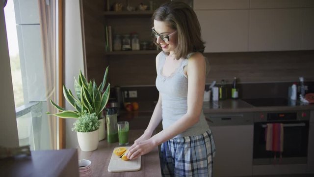 Young beautiful female in pajamas and eyeglasses cutting orange standing in kitchen.