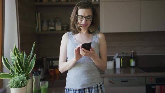 Crop young smiling female in eyeglasses using smartphone standing in kitchen.