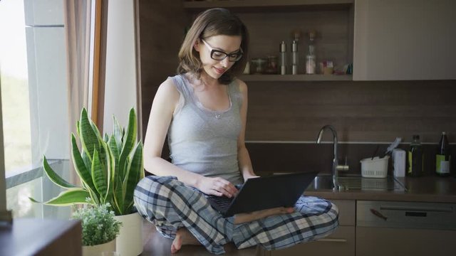 Attractive female in pajamas and eyeglasses sitting on top of table in kitchen and using laptop. 