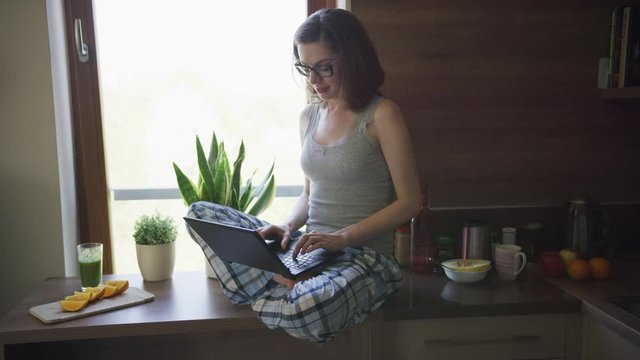 Young female in pajamas and eyeglasses sitting on top of table in kitchen and using laptop. 