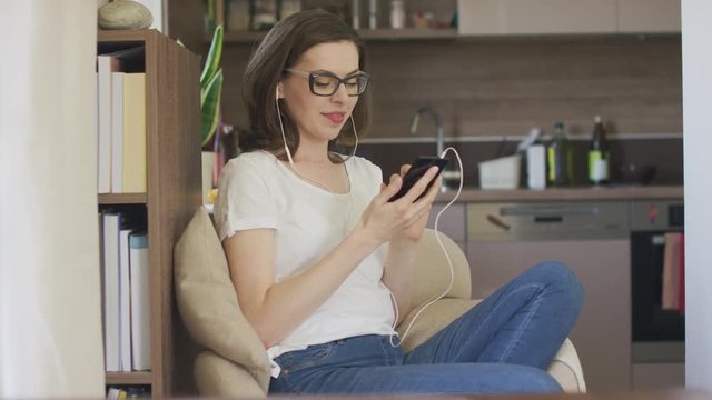 Young beautiful female in eyeglasses sitting in comfortable armchair at home and using smartphone with earphones. 