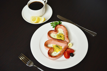 Scrambled eggs in the form of heart on a white plate with sausages, tomatoes, greens and coffee with lemon.