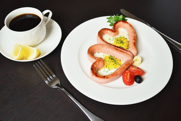 Scrambled eggs in the form of heart on a white plate with sausages, tomatoes, greens and coffee with lemon.