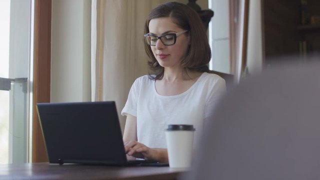 Young beautiful female in eyeglasses sitting at home before laptop and using smartphone. 