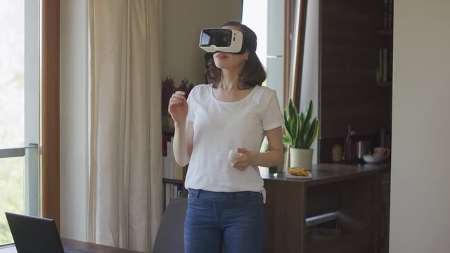 Young female standing in virtual reality headset and holding remote controller at home.