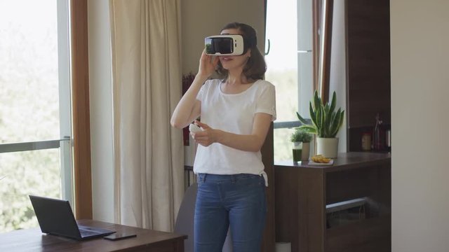Young female standing in virtual reality headset at home and pushing button on remote controller. 