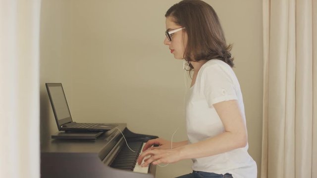 Side view of attractive female in eyeglasses and earphones playing digital piano looking at laptop screen.