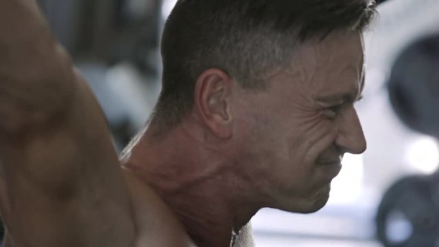 Shirtless middle-aged man working out at the gym and performing squat exercises when holding heavy barbell overhead