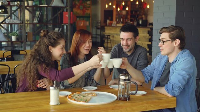 Friendly team is celebrating successful end of project in cafe with drinks and pizza, they are talking and clanging cups drinking tea. Eating out and friendship concept.