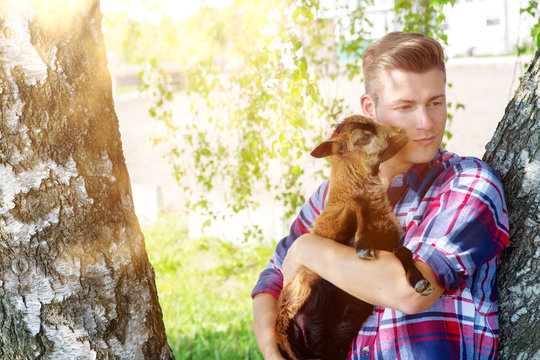 Blond Man Holding A Baby Goat Next To A Tree