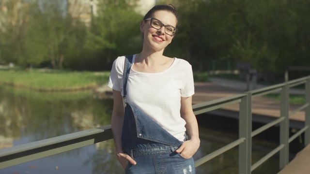 Attractive young female in eyeglasses standing leaning on handrail above water in park and looking at camera.