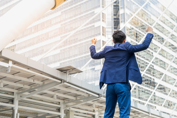 Fototapeta premium Back of businessman stand and raising up his hands to cheerful and celebrated his successful in career and mission. concept of winner and compettition with copy space for text.