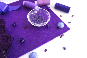 Fototapeta na wymiar A set of materials for needlework in violet color on a white background. Beads, threads, a satin ribbon and wool for knitting.
