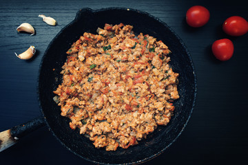 Fried ground meat with vegetables in an old frying pan on a dark background, garlic and tomatoes