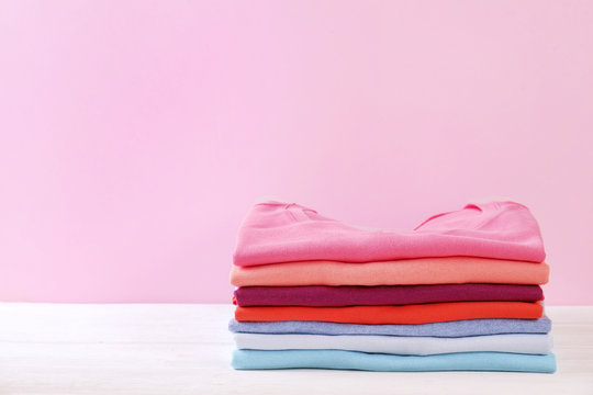 Stack of colorful perfectly folded sweaters on white wood texture table. Pile of different pastel color shirts and sweaters on wooden table, pale pink wall background. Close up, top view, copy space.