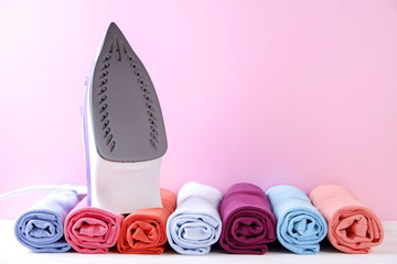 Stack of clean freshly laundered, neatly rolled clothes and electric iron on pressboard. Pile of t-shirts rolls on white wooden ironing board, pink wall background. Copy space, close up, top view.