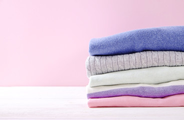 Fototapeta na wymiar Stack of colorful perfectly folded sweaters on white wood texture table. Pile of different pastel color shirts and sweaters on wooden table, pale pink wall background. Close up, top view, copy space.