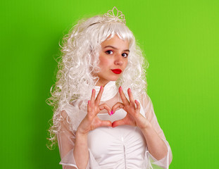 Charming brown-eyed girl in long blonde hair wig making love heart gesture on green bright background