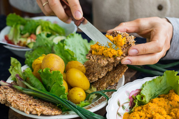 Woman hands spreads chickpea hummus on piece of whole wheat bread. Vegetarian lunch with backed new potatoes, salad, onion. Raw vegan vegetarian healthy food