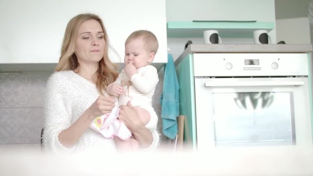 Mom with baby going in white kitchen. Beauty woman with little child on hand. Mother with baby on hands licks finger and go away
