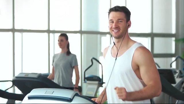 Smiling Athletic Man Running On Treadmill. Slow Motion Handsome Guy Doing Workout At Gym And Listening Music. Sport And Healthy Lifestyle.