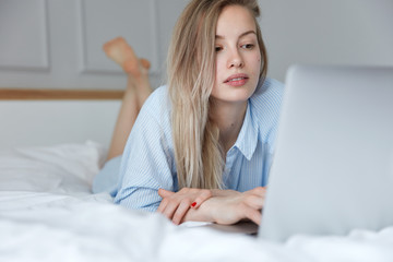 Concentrated European female bloger in striped casual night shirt, types article and surfes her blog before sleep, poses on comfortable bed against blurred bedroom interior, keeps feet in air.